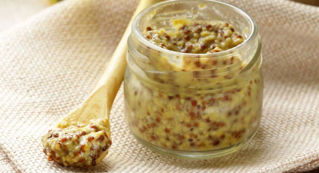 A glass jar of whole grain mustard with seeds visible, next to a wooden spoon also covered in mustard, placed on a beige textured cloth.