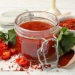 A jar of red chili jam surrounded by sliced red chilies, a bowl of red chili flakes, parsley leaves, a garlic bulb, and a wooden scoop of chili powder on a wooden table.