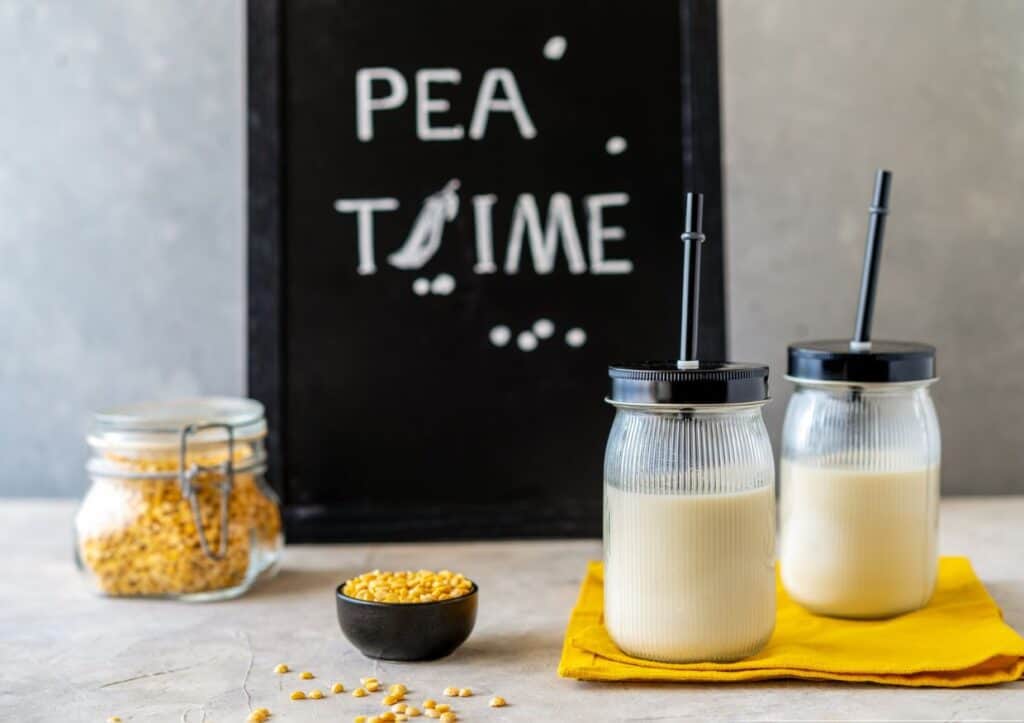 Two jars of pea milk with straws on a cloth beside a chalkboard reading "Pea Time" and jars of dry peas.