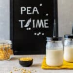 Two jars of pea milk with straws on a cloth beside a chalkboard reading "Pea Time" and jars of dry peas.