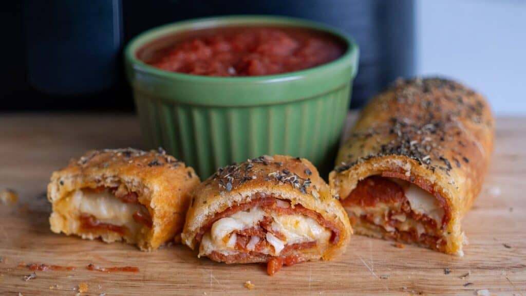 Close-up of sliced pizza rolls with melted cheese and pepperoni on a wooden board, with a green bowl of red dipping sauce in the background.