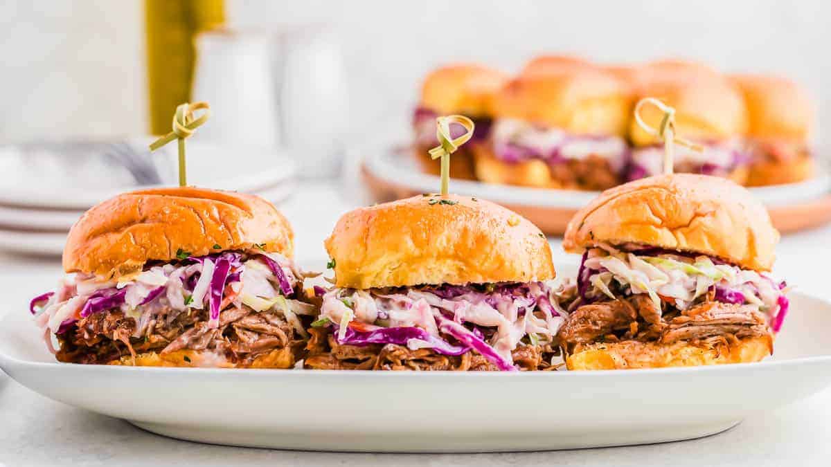 Three pulled pork sliders topped with coleslaw on a white serving plate, with another plate of sliders blurred in the background. Each slider is held together with a bamboo skewer.