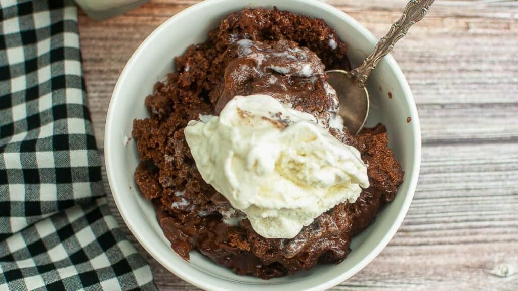 A bowl of chocolate pudding cake topped with melting vanilla ice cream and a spoon, placed on a wooden surface beside a checkered cloth.
