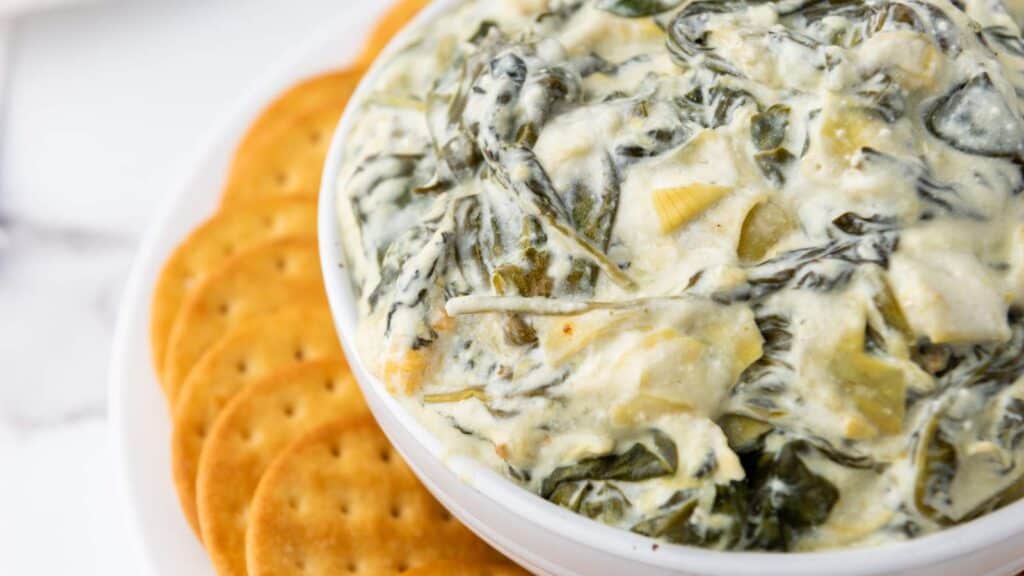 A bowl of creamy spinach artichoke dip is placed next to a stack of round crackers.