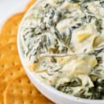 A bowl of creamy spinach artichoke dip is placed next to a stack of round crackers.