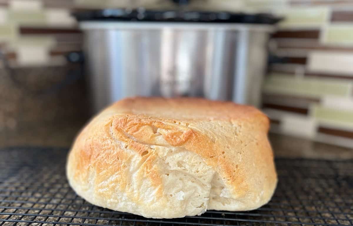A loaf of fresh bread cools on a black wire rack in front of a slow cooker, with a tiled kitchen backsplash in the background.