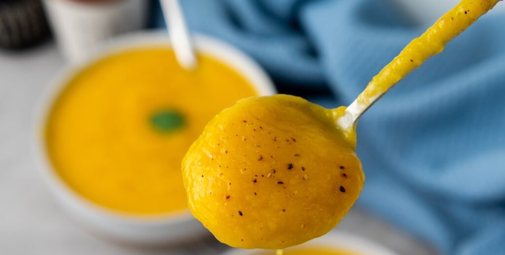 A spoon holding a scoop of creamy yellow soup, possibly squash or pumpkin, with visible pepper flecks. A bowl of the same soup and a blue cloth are blurred in the background.