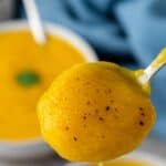 A spoon holding a scoop of creamy yellow soup, possibly squash or pumpkin, with visible pepper flecks. A bowl of the same soup and a blue cloth are blurred in the background.