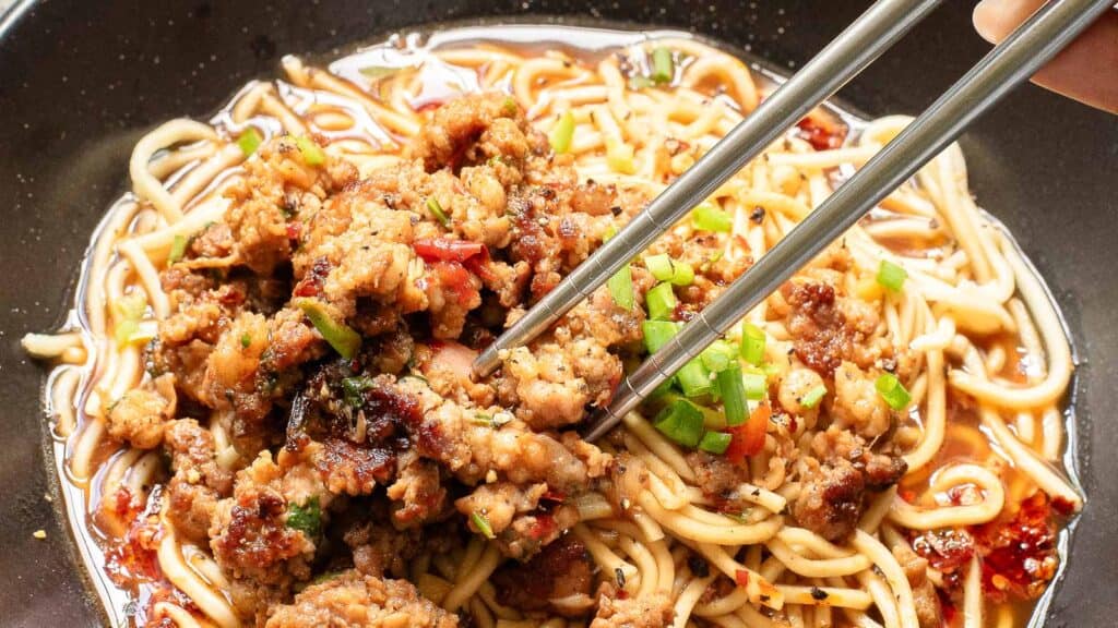 Plate of noodles topped with minced meat and vegetables, with chopsticks picking up a portion.