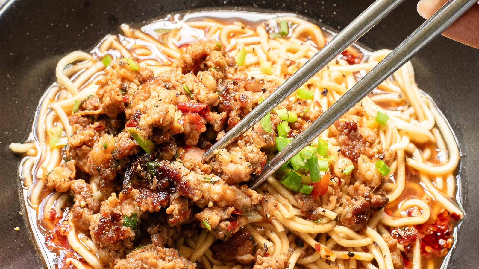 A bowl of tantanmen noodles topped with minced meat and vegetables is being picked up with chopsticks. The dish includes colorful ingredients like red and green peppers, all served in a rich, savory sauce on a dark plate.