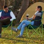 A man and woman sit on camping chairs, holding wine glasses, in front of a tent and a lit fire pit.