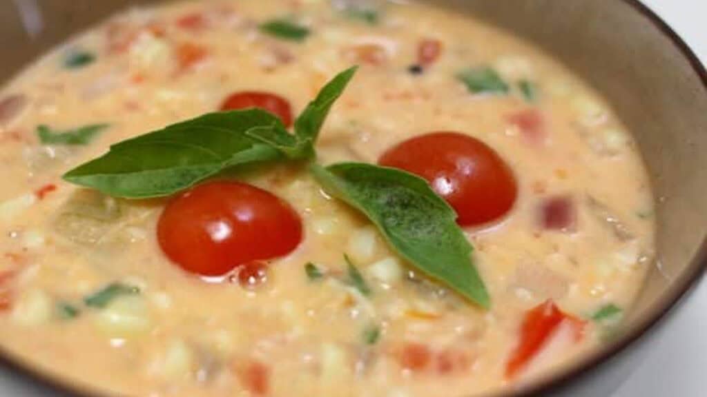 A bowl of creamy soup with cherry tomatoes, diced vegetables, and fresh basil leaves.