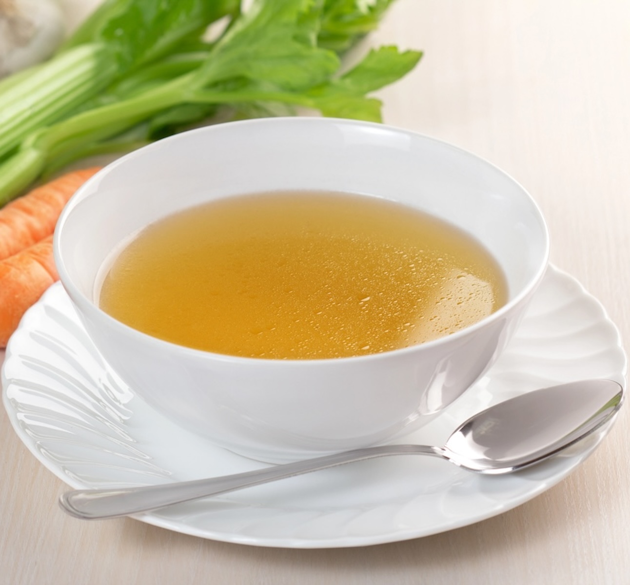 A bowl of clear broth on a white plate with a spoon, surrounded by fresh vegetables like celery and carrot.