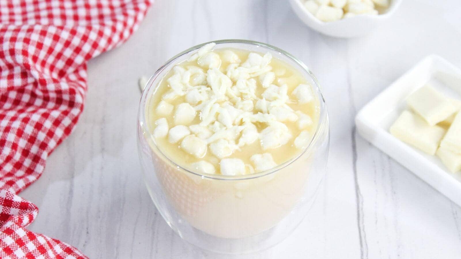 Glass of creamy white dessert topped with white chocolate shavings. Red gingham cloth and a small bowl with more toppings are nearby on a white surface.
