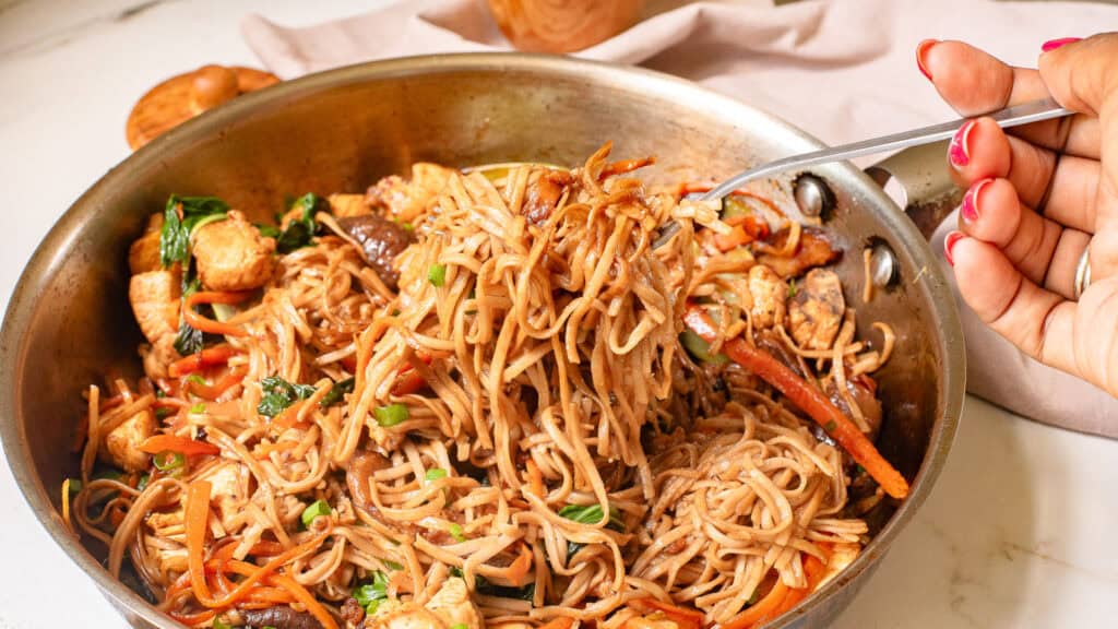 A hand holding a fork with cooked noodles, vegetables, and chunks of chicken in a pan.