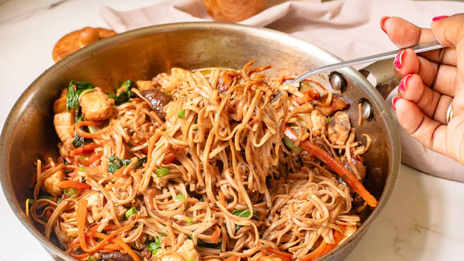 A fork lifts noodles from a white bowl filled with stir-fried noodles and vegetables. A skillet with more noodles is in the background on a marble surface.