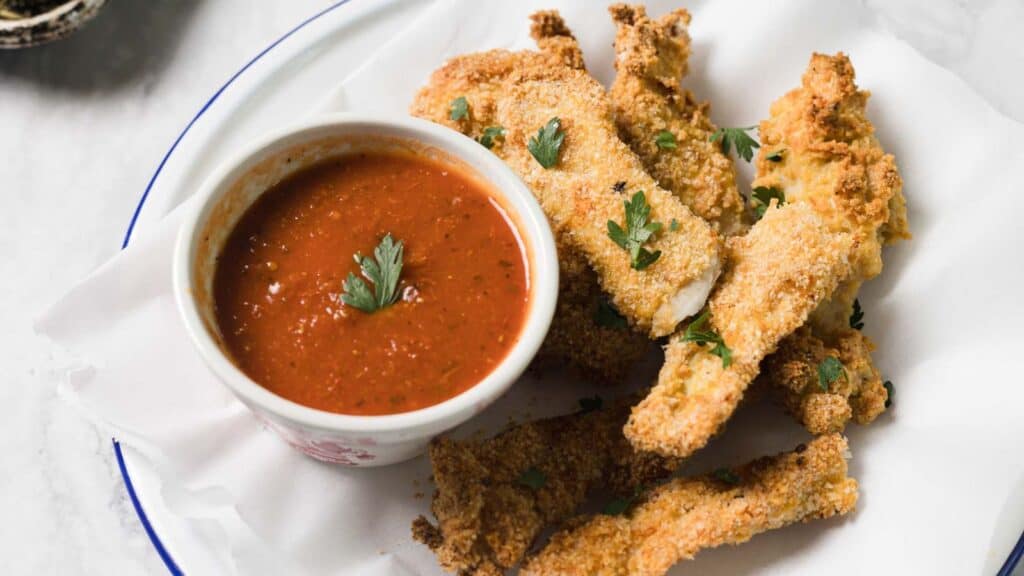A plate of breaded fried chicken strips with parsley, served with a bowl of red dipping sauce, makes for one of those easy dinners that's both satisfying and delicious.