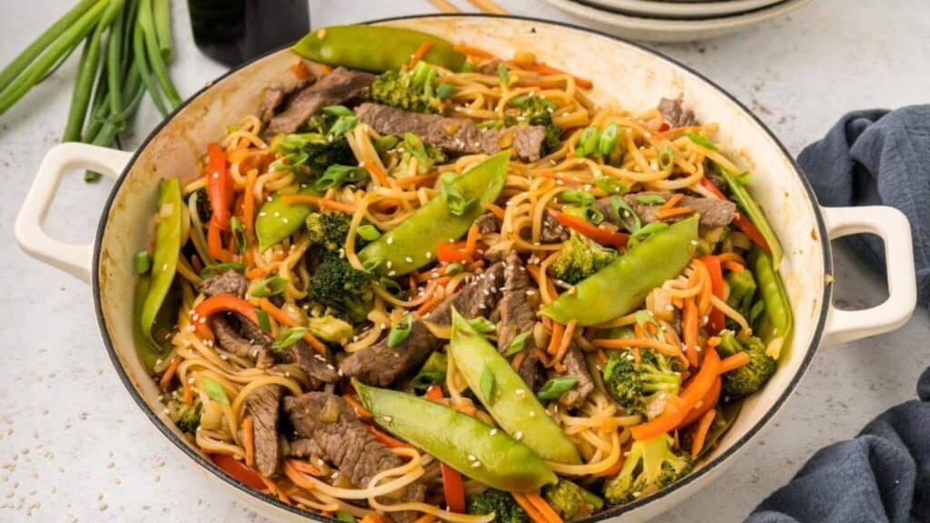 A pan filled with stir-fried noodles, beef slices, broccoli, carrots, snap peas, and sprinkled with sesame seeds.