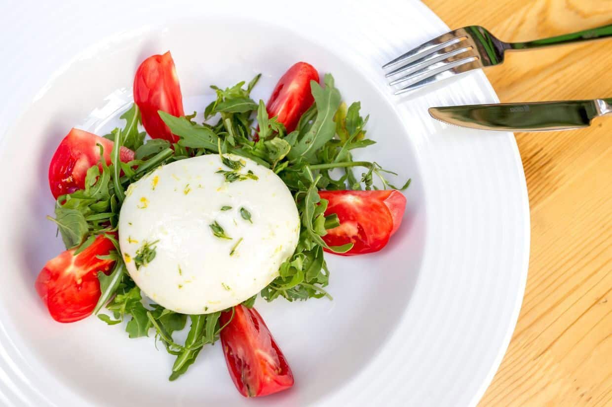A plate with a round burrata cheese on a bed of arugula, surrounded by tomato wedges. A fork and knife are placed beside the plate.