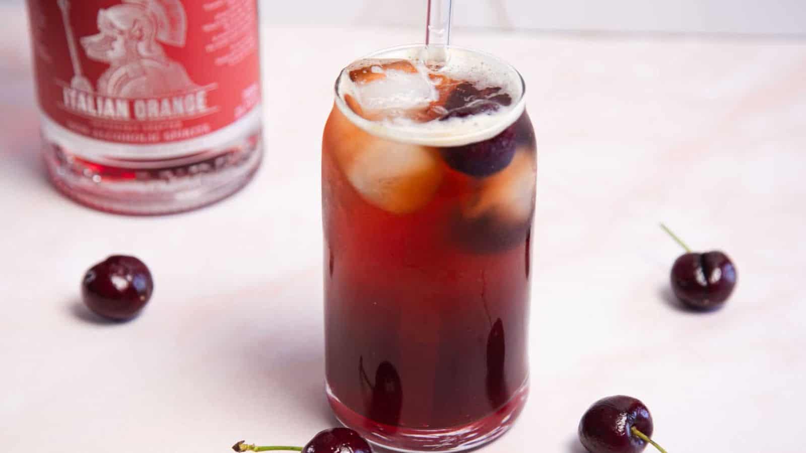 A tall, clear glass filled with a dark red drink, ice cubes, and garnished with cherries. A clear straw is inserted into the drink. Surrounding the glass are fresh cherries, with a bottle labeled 