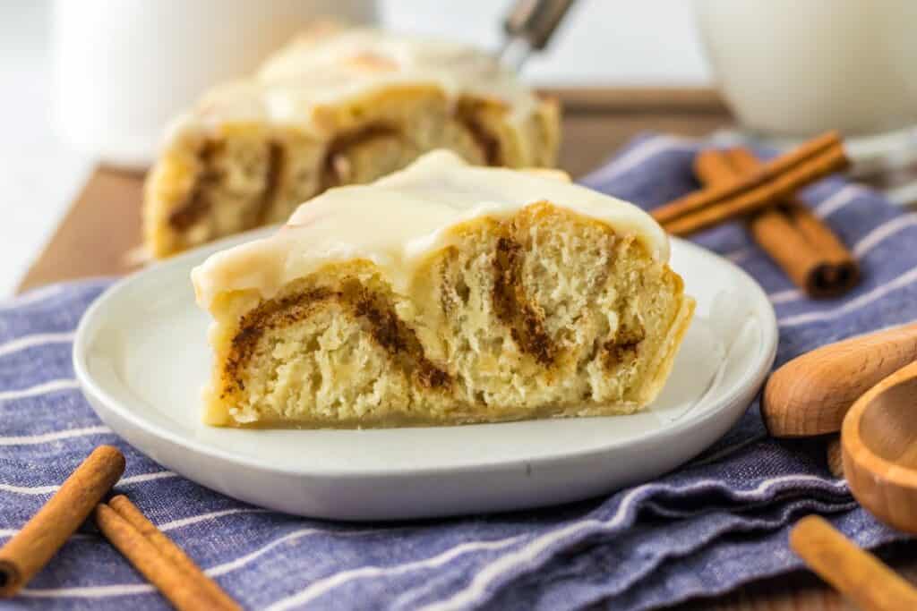 A slice of cinnamon roll with icing on a white plate, placed on a striped cloth. Cinnamon sticks are nearby.