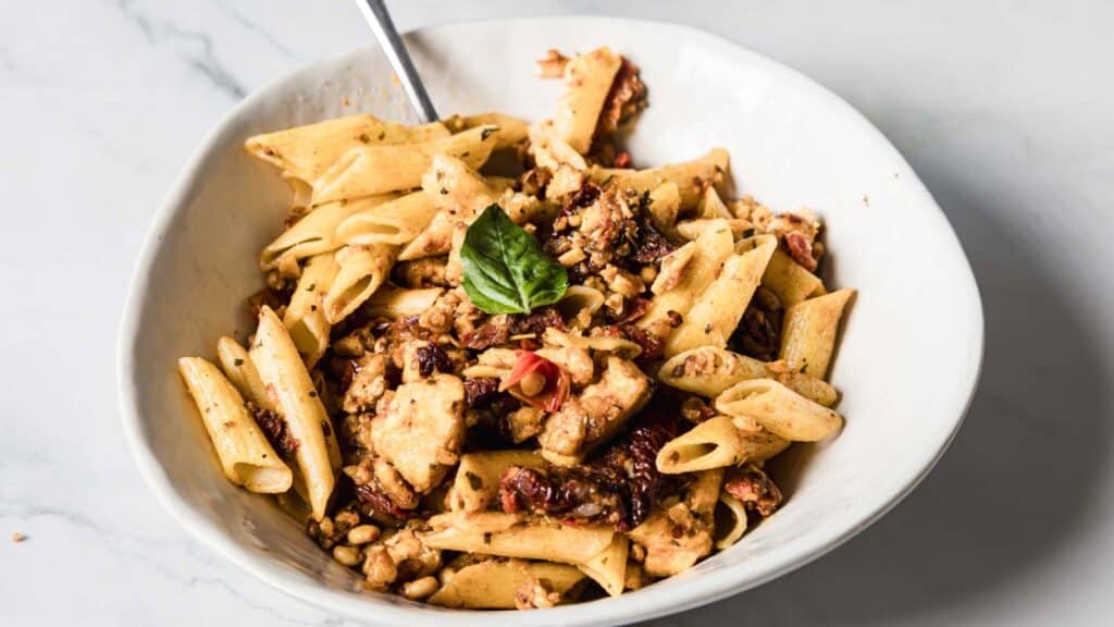 A white bowl filled with penne pasta mixed with chunks of chicken, sun-dried tomatoes, and garnished with fresh basil leaves. A fork is placed in the bowl, and a small dish is visible in the background.
