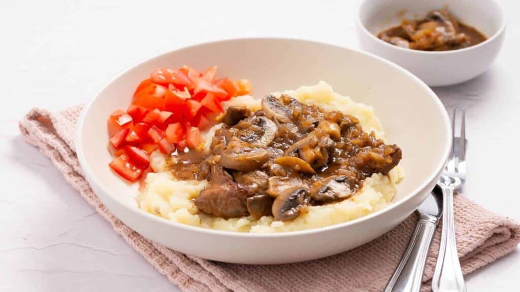 A bowl of mashed potatoes topped with beef and mushroom stew, garnished with diced tomatoes. A small bowl of stew is in the background beside utensils on a napkin.