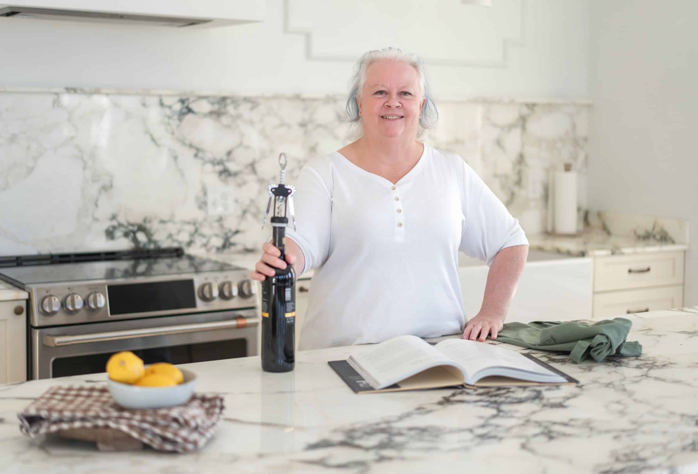A woman in a white top stands in a kitchen with marble countertops, holding a bottle opener on a wine bottle next to an open book, two lemons in a bowl, and a folded green cloth.