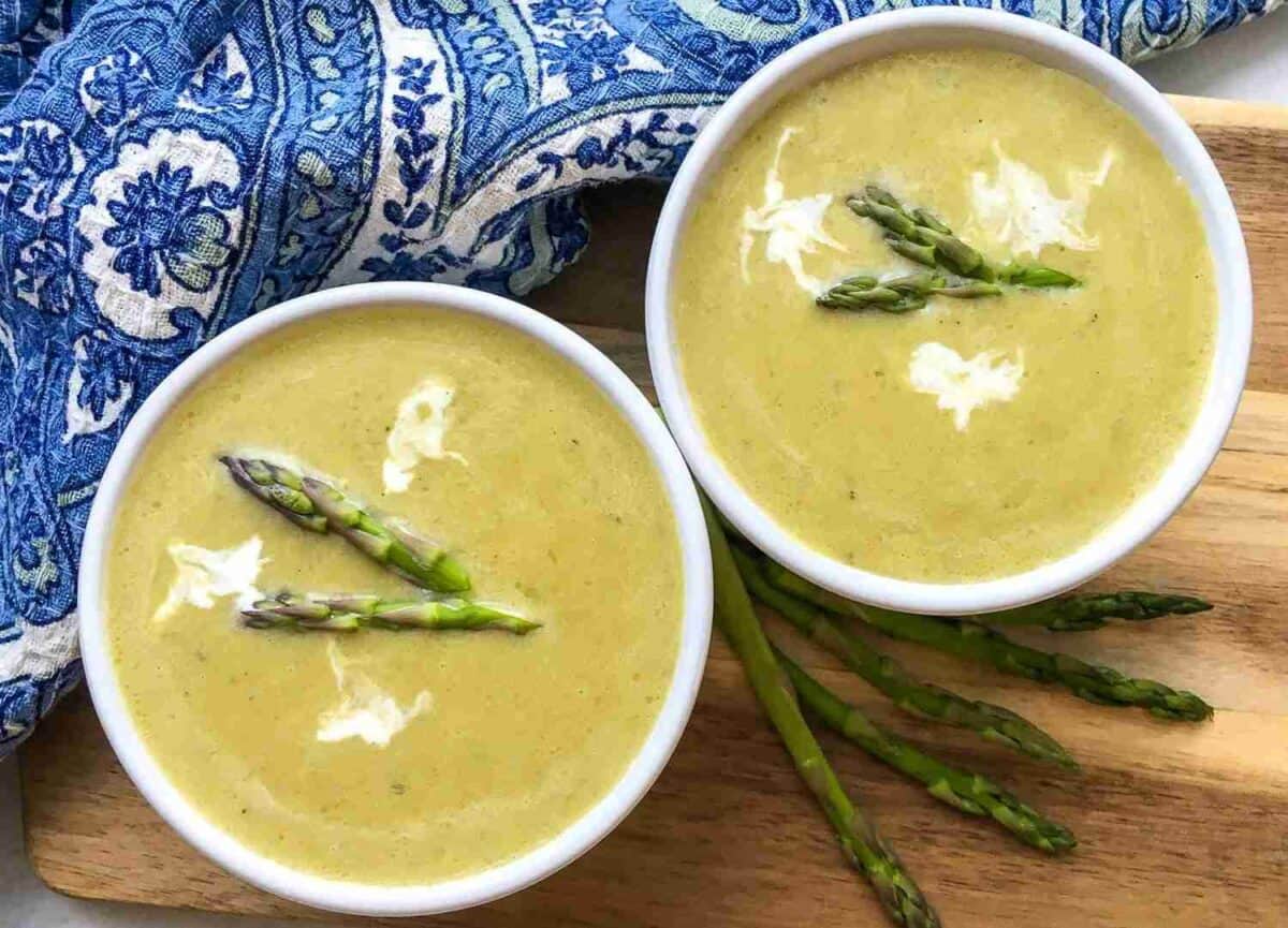 Two bowls of asparagus soup with cream swirls and asparagus tips, placed on a wooden surface. A blue patterned cloth is partially visible in the background.
