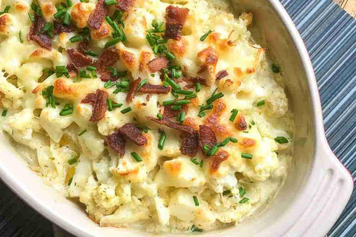 Baked cauliflower casserole topped with melted cheese, crispy bacon bits, and chopped chives in an oval dish on a striped surface.
