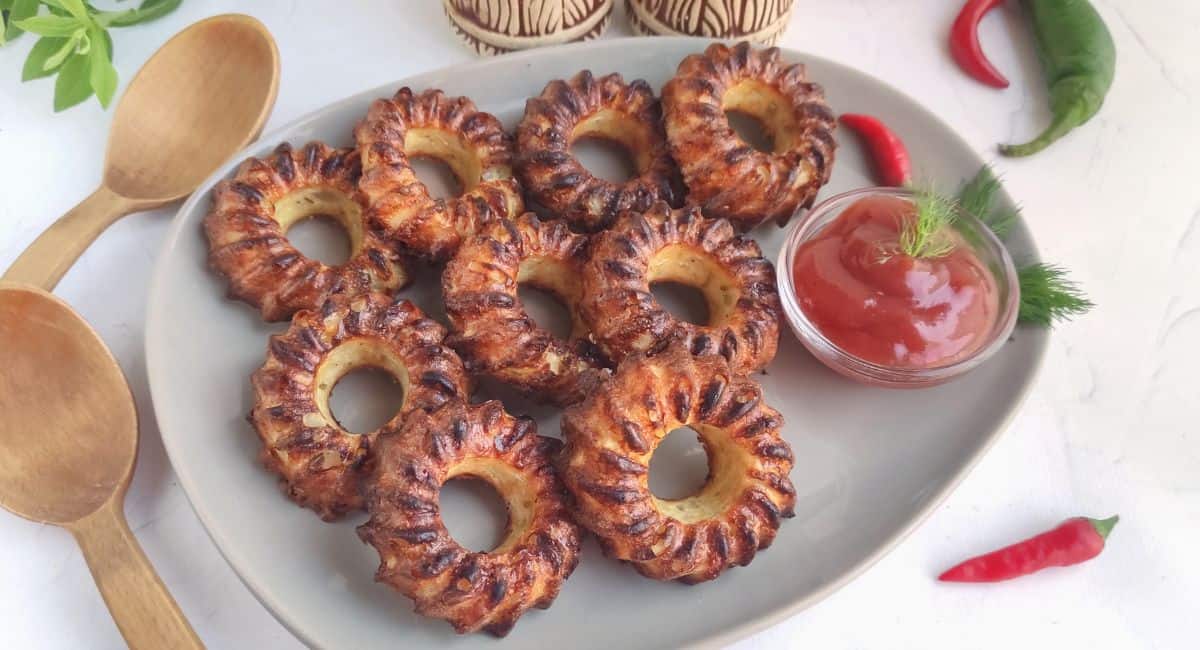 A plate of crispy ring-shaped pastries with a small bowl of red dip, garnished with dill. Two wooden spoons and chili peppers are placed nearby.