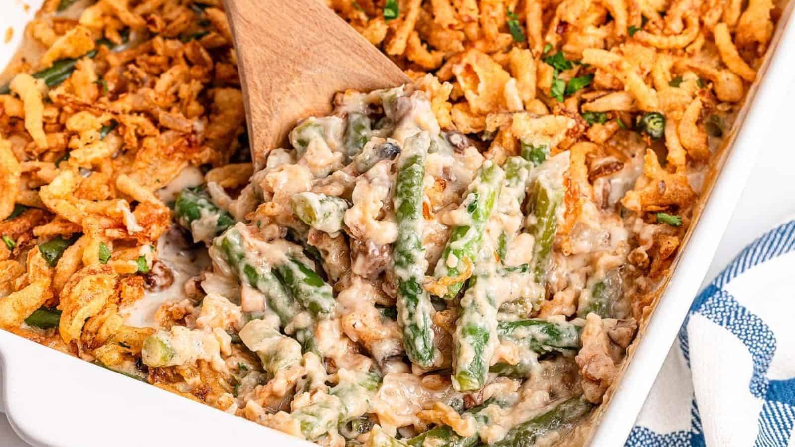 Green bean casserole in a white baking dish, topped with crispy fried onions, with a wooden spoon in the dish.