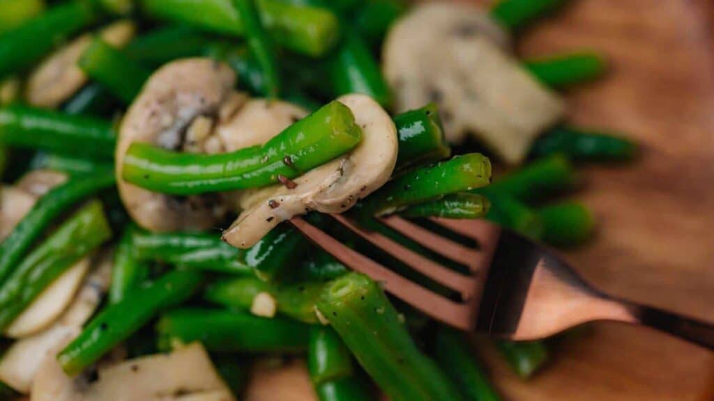 A close-up of a fork holding saut&eacute;ed green beans and mushrooms.