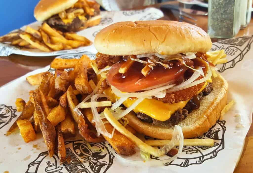 A cheeseburger topped with onion rings, tomato, and sauce is served on a plate alongside a generous helping of fries—perfect cruise food for when you're relaxing by the pool.