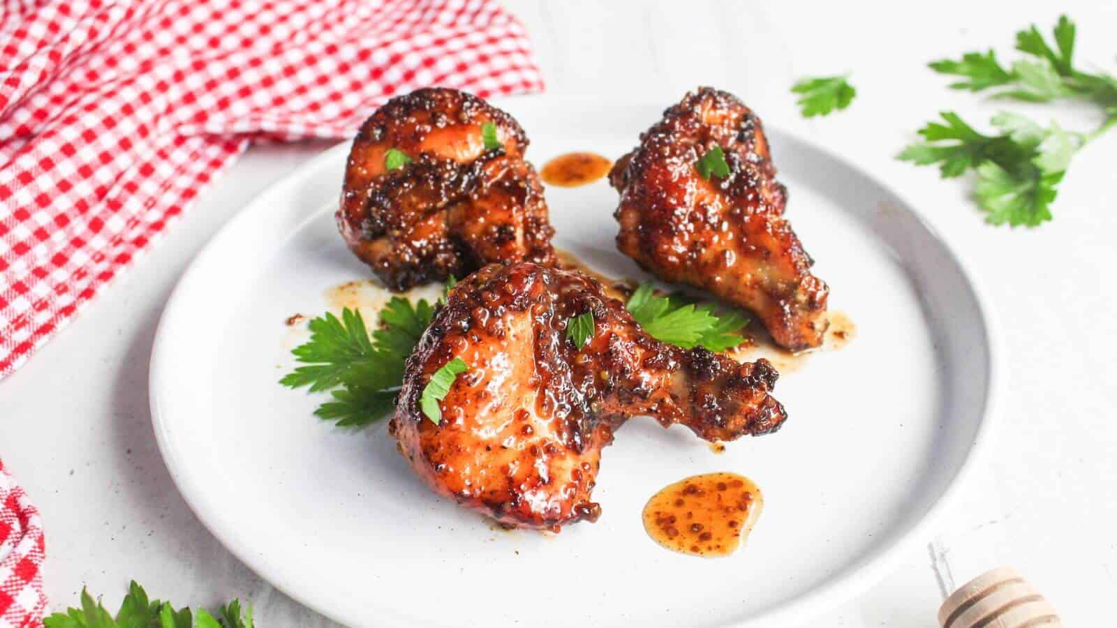 Three glazed chicken wing pieces garnished with parsley are arranged on a white plate. A red and white checkered cloth is visible beside the plate.