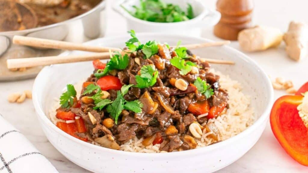 A bowl of rice topped with beef stir-fry, red bell peppers, peanuts, and cilantro. Chopsticks rest on the bowl. Additional ingredients are visible in the background.