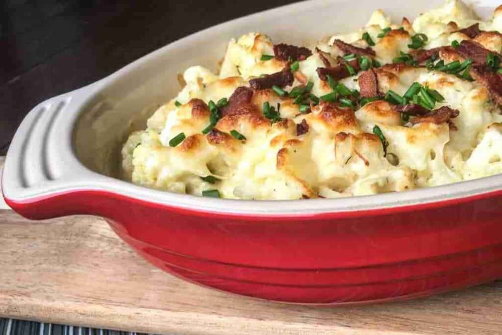 Baked cauliflower topped with melted cheese, crispy bacon, and chopped chives in a red dish.
