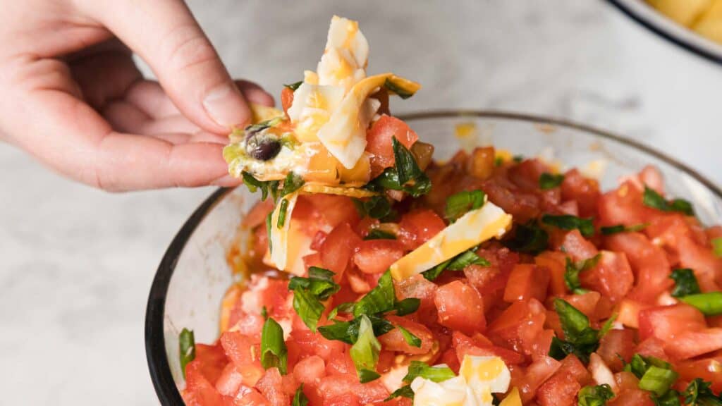A hand holding a cracker topped with chopped tomatoes, cheese, black olives, and green peppers over a bowl of similar ingredients.