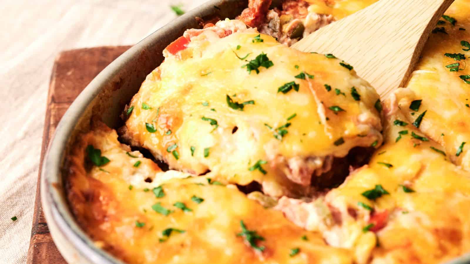 A cheesy casserole with chopped herbs on top, being served with a wooden spatula, in a round baking dish.