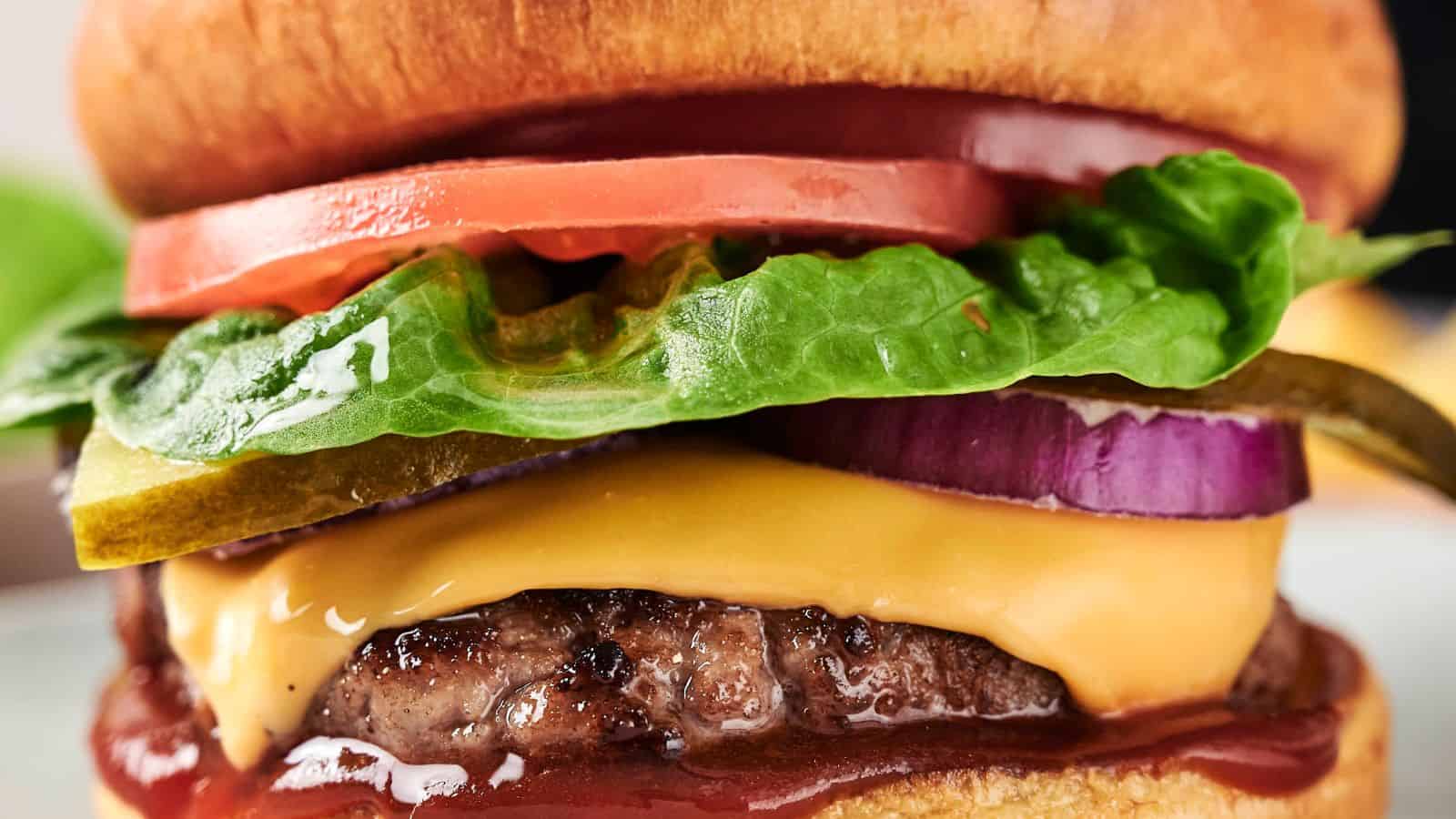 Close-up of a cheeseburger with lettuce, tomato, pickles, red onion, and a beef patty topped with melted cheese and ketchup in a bun.
