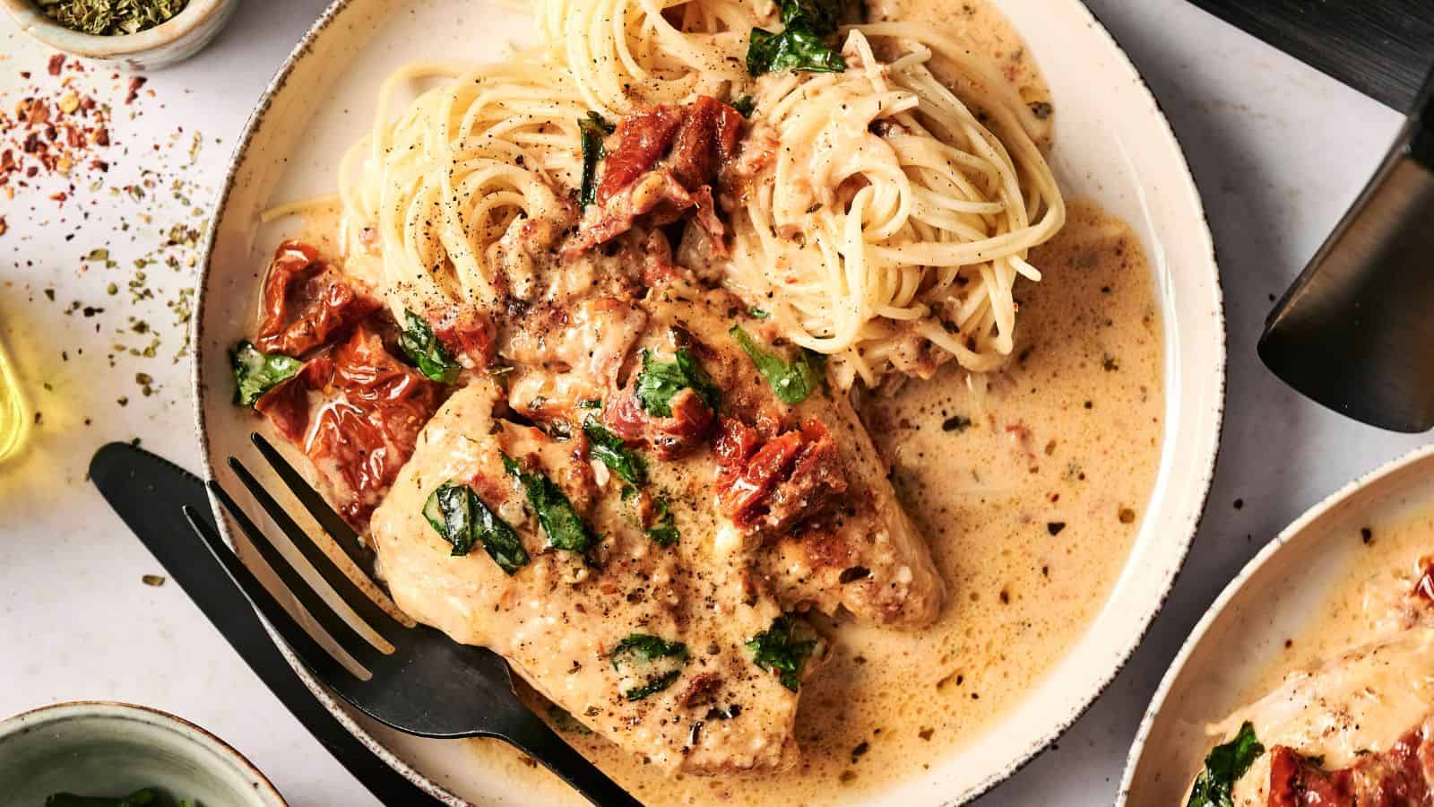 Plate of creamy chicken with sun-dried tomatoes and basil, served with spaghetti. A fork and knife rest on the plate.