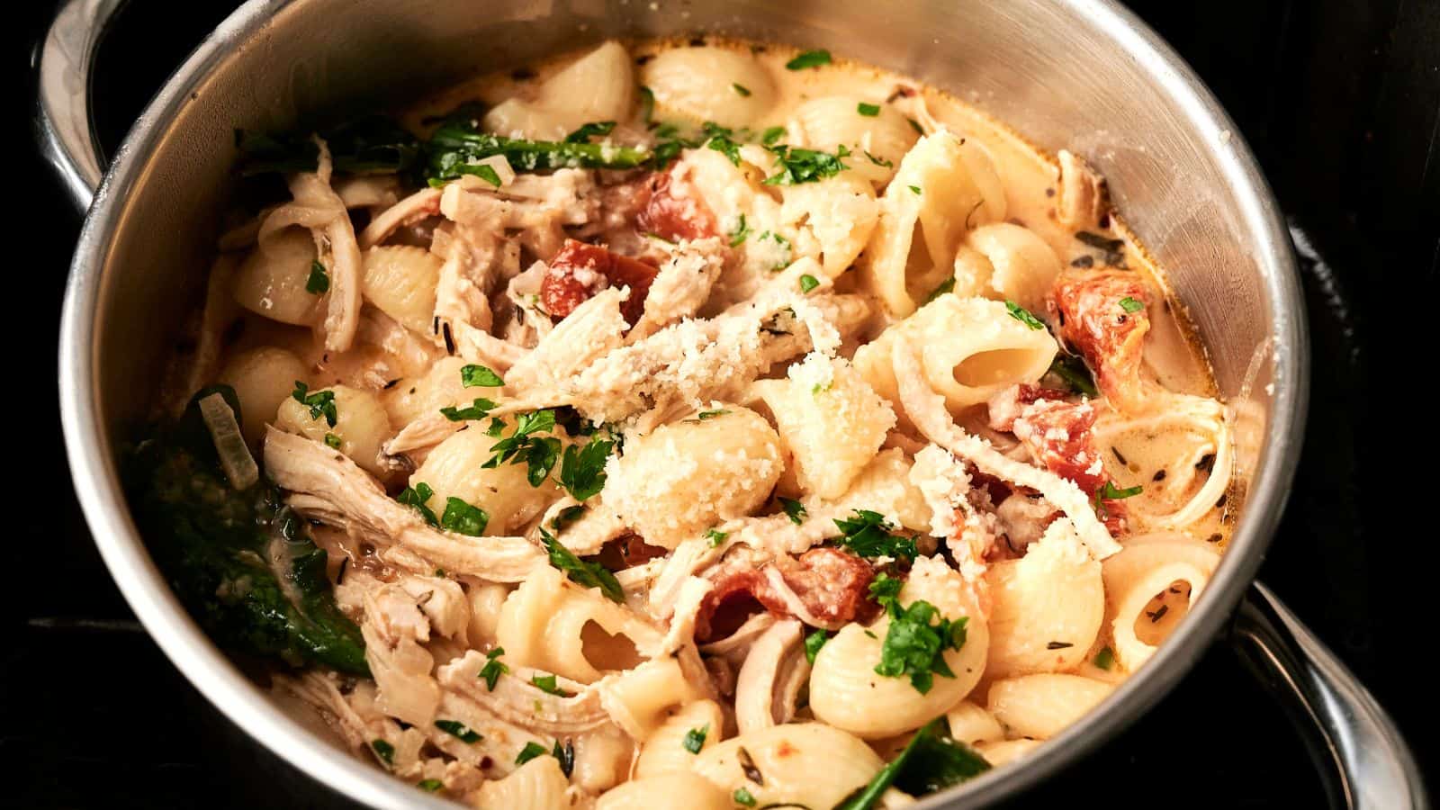 A pot of pasta with shredded chicken, spinach, tomatoes, and grated cheese, garnished with chopped parsley.