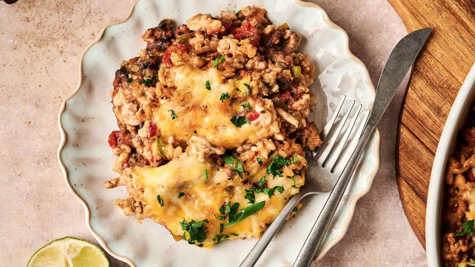A plate with a serving of cheesy casserole containing ground meat, beans, and chopped herbs, accompanied by a fork and knife. A lime wedge is partially visible on the side.