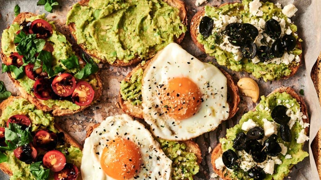 Assorted avocado toasts with toppings: cherry tomatoes, parsley, fried eggs, spices, feta cheese, and black olives on a parchment-lined surface.
