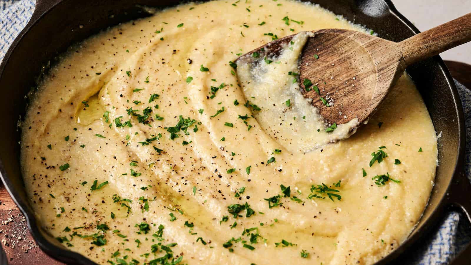 Creamy polenta in a skillet, topped with chopped herbs and black pepper, and stirred with a wooden spoon.