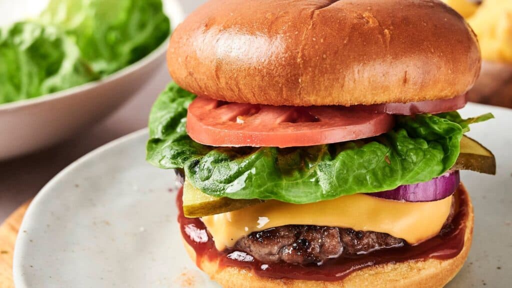 A cheeseburger with lettuce, tomato, pickles, onion, and sauce on a plate, with a bowl of lettuce in the background.