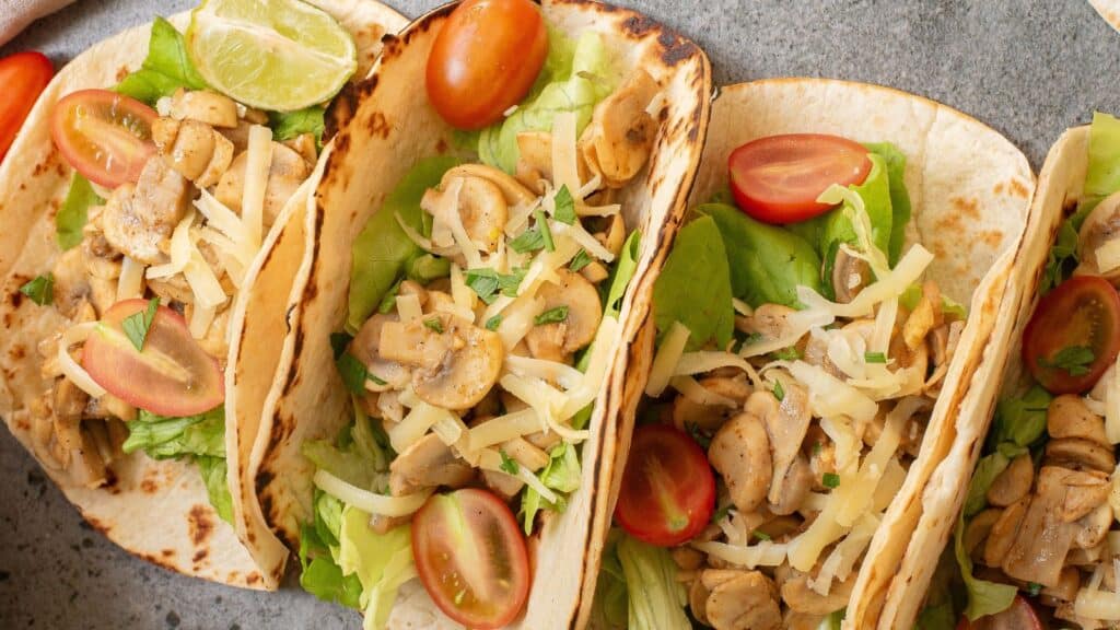 Three tacos filled with lettuce, sliced mushrooms, shredded cheese, and halved cherry tomatoes, served with a lime wedge on the side.