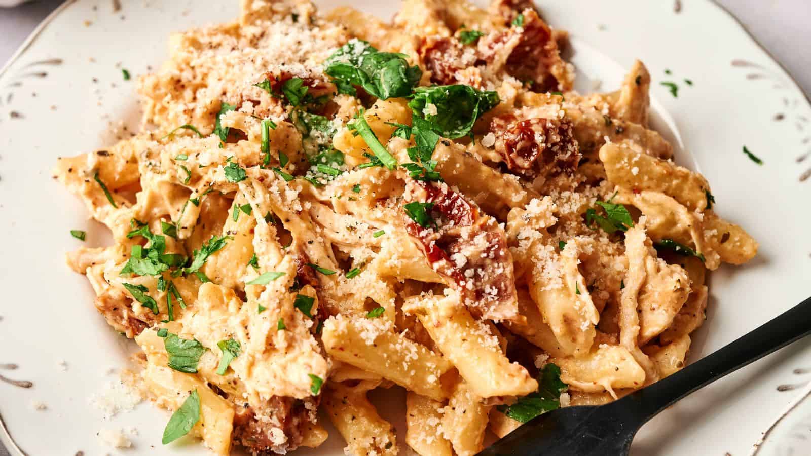 A plate of creamy penne pasta garnished with chopped parsley and grated cheese, featuring pieces of sun-dried tomatoes. A black fork is resting on the plate.