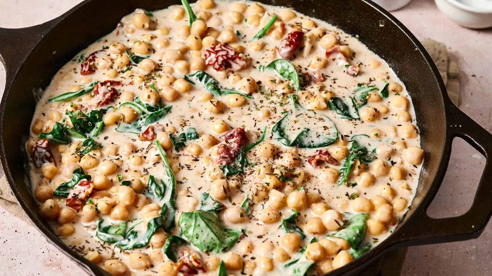 Skillet with creamy chickpeas, spinach, and sun-dried tomatoes, garnished with herbs.