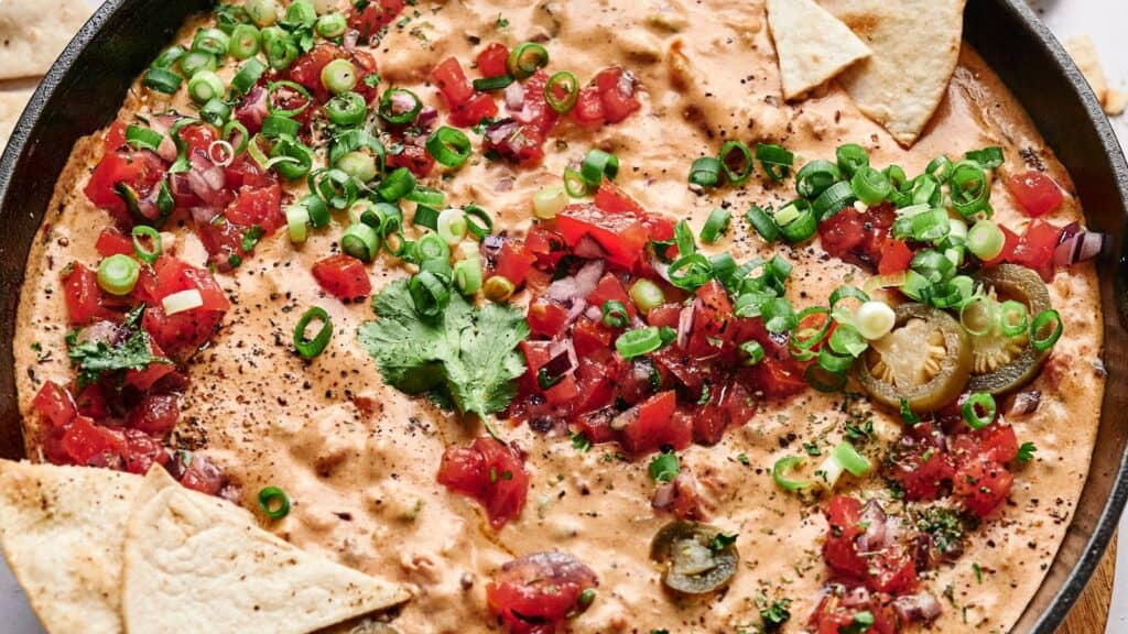 A skillet filled with creamy cheese dip topped with diced tomatoes, jalape&ntilde;os, green onions, cilantro, and surrounded by tortilla chips.